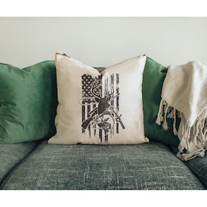 May include: A decorative throw pillow with a white background and a brown silhouette of a deer with a fishing rod and fish. The deer is standing in front of an American flag.