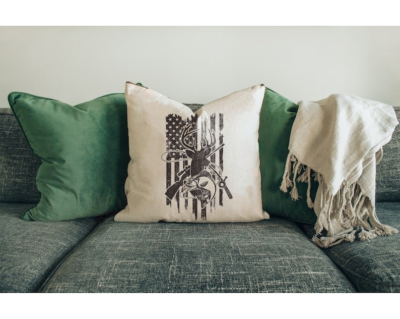 May include: A decorative throw pillow with a white background and a brown silhouette of a deer with a fishing rod and fish. The deer is standing in front of an American flag.