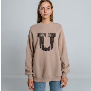 May include: A beige sweatshirt with a large black letter "U" printed on the front. The letter has a distressed, vintage look.