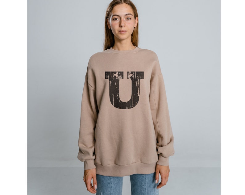 May include: A beige sweatshirt with a large black letter "U" printed on the front. The letter has a distressed, vintage look.