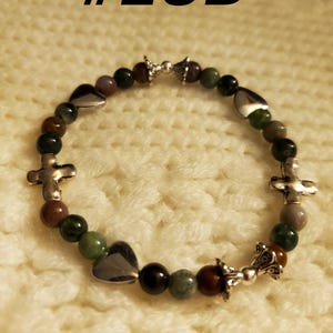 May include: A green, brown, and purple stone bracelet with silver heart and cross charms. The bracelet has a silver clasp.