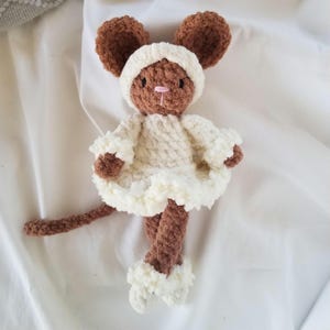 May include: A brown crocheted mouse plush toy featuring large rounded ears, a pink stitched nose, and dark eyes. The mouse wears a cream-colored crocheted bonnet, a matching ruffled dress with flared sleeves, and small cream-colored booties on its feet.