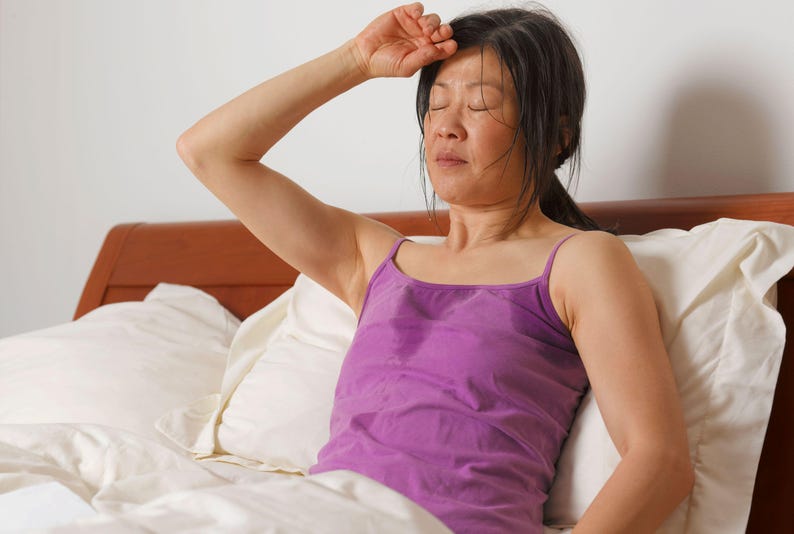 May include: A woman in a purple tank top is lying in bed with her hand on her forehead. She appears to be sweating.