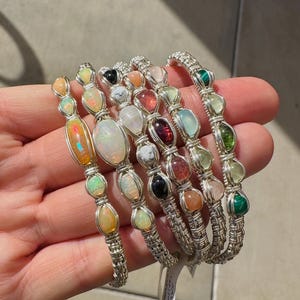 May include: A selection of handcrafted bracelets, each featuring a woven silver wire design and a variety of gemstones. The bracelets include opal, quartz, and malachite. The colourful stones are set in a silver wire frame. The bracelets are displayed on a hand.