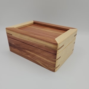 May include: A rectangular wooden box with a lid. The box is constructed from two types of wood: reddish-brown and light-colored. The corners are dovetailed, and the lid sits flush with the box. The box is empty.
