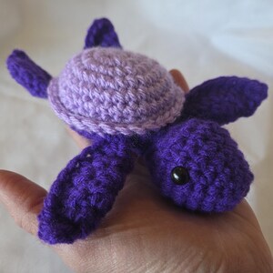 Puede incluir: Una tortuga de crochet de color morado y lavanda con ojos negros. La tortuga está hecha de hilo suave y es una gran adición a la colección de juguetes de cualquier niño.