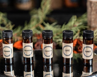 May include: Six small brown glass bottles with labels on a silver tray. The labels are for "Abundance", "Protection", "Money", "Love", and "Abundant". The bottles are surrounded by dried orange flower petals and green pine branches.