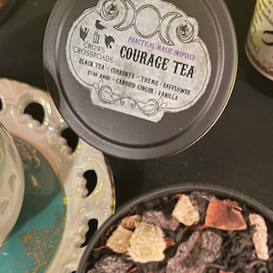 May include: A black tin with a white label that reads "CROWS CROSSROADS PRACTICAL MAGIC INSPIRED COURAGE TEA BLACK TEA - CURRENTS - THYME - SAFFLOWER STAR ANISE - CANDIED GINGER - VANILLA". The tin is filled with loose leaf tea.