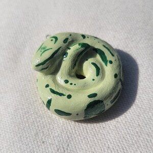 May include: A small, green and white ceramic snake charm. The snake is curled up in a circle and has a green and white pattern.
