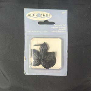 May include: A black decorative leaf pull for window shades, packaged in a clear plastic blister pack. The package includes a screw for installation. The product is labeled "ACCENTS on SHADES".