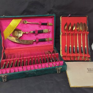 以下が含まれることがあります： 緑色のベルベットケースにピンクの裏地が付いた、ヴィンテージのスターハウスカトラリーセット。フォーク、ナイフ、スプーン、サービングスプーンなど、黒いハンドルが付いた金色の道具が含まれています。ケースは開いており、カトラリーの配置が明らかになっています。