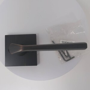 Puede incluir: Una manija de puerta negra con una placa posterior cuadrada y un diseño de palanca. La manija es de metal y tiene un acabado cepillado. La manija viene con una pequeña bolsa de tornillos etiquetada "5049602".