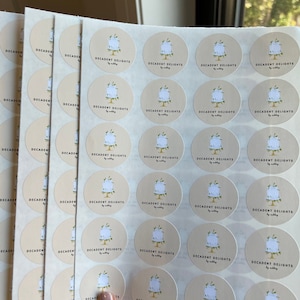 Peut inclure: Une pile de feuilles de papier autocollant blanc avec des autocollants ronds beiges imprimés avec le texte "Decadent Delights by Sally". Chaque autocollant présente une illustration stylisée d'un gâteau avec des feuilles vertes.