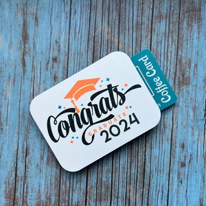 May include: A white gift card with the text "Congrats Graduate 2024" in black and orange lettering. A graduation cap icon is above the text. A blue gift card with the text "Coffee Card" is attached to the right side of the white card.