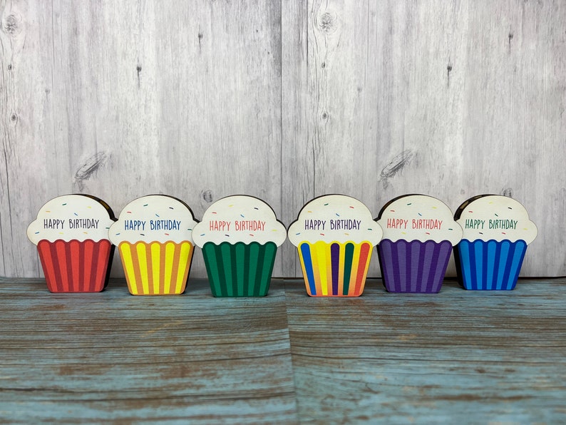 May include: Six wooden cupcake decorations with the words "Happy Birthday" on each. The cupcakes are in different colors: red, yellow, green, purple, blue, and orange.