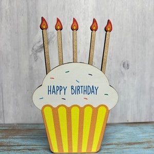 May include: A wooden cupcake with yellow frosting and five red and orange candles. The cupcake has the words "Happy Birthday" written on it.