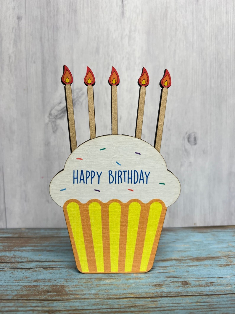 May include: A wooden cupcake with yellow frosting and five red and orange candles. The cupcake has the words "Happy Birthday" written on it.