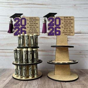 May include: Two graduation-themed money holders. One is filled with rolled dollar bills. Each has a wooden sign with the year "2026" in purple, a graduation cap, and text that reads "Behind you all your memories... Congratulations Graduate!"