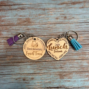 May include: Two wooden keychains with silver keyrings and colorful tassels. One is round and says "Bland Elementary Lunch Team." The other is heart-shaped and says "Laura Lunch Team." Both have engraved text.