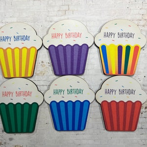 May include: Six wooden cupcake-shaped ornaments with the text "Happy Birthday" on each. The cupcakes are decorated with colorful sprinkles and have different colored frosting: yellow, purple, rainbow, green, blue, and red.