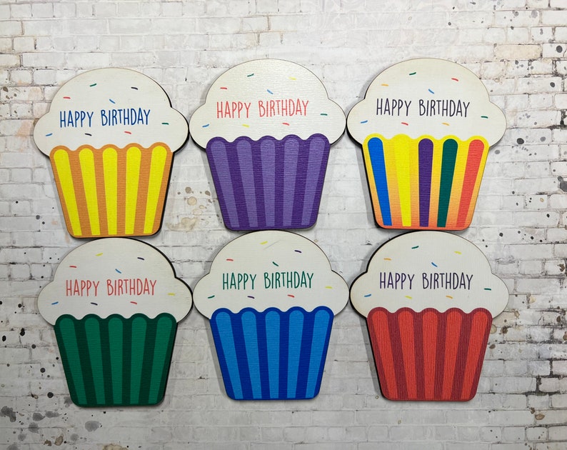 May include: Six wooden cupcake-shaped ornaments with the text "Happy Birthday" on each. The cupcakes are decorated with colorful sprinkles and have different colored frosting: yellow, purple, rainbow, green, blue, and red.