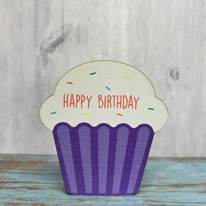 May include: A wooden cupcake-shaped sign with the words "Happy Birthday" in red lettering. The cupcake is purple with white frosting and colorful sprinkles.