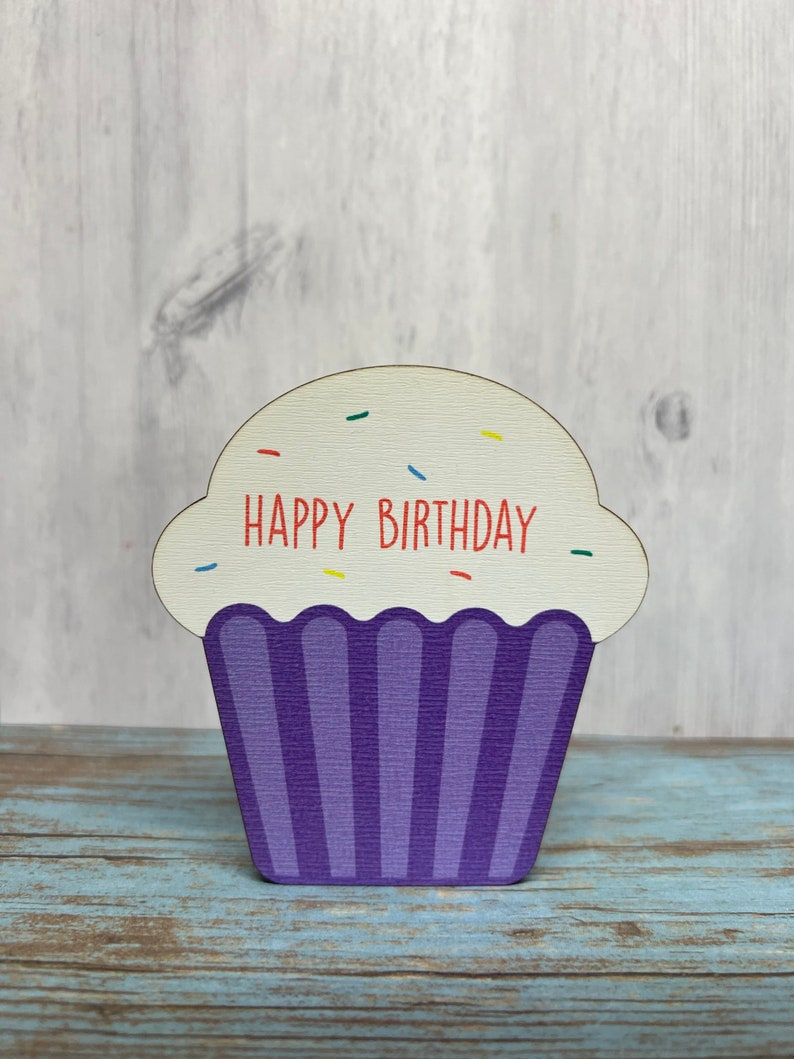 May include: A wooden cupcake-shaped sign with the words "Happy Birthday" in red lettering. The cupcake is purple with white frosting and colorful sprinkles.
