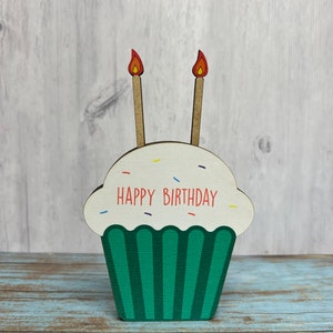 May include: A wooden cupcake with green frosting and two candles with red flames. The cupcake has white sprinkles and the text "HAPPY BIRTHDAY" in red letters.