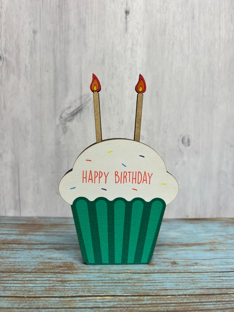 May include: A wooden cupcake with green frosting and two candles with red flames. The cupcake has white sprinkles and the text "HAPPY BIRTHDAY" in red letters.