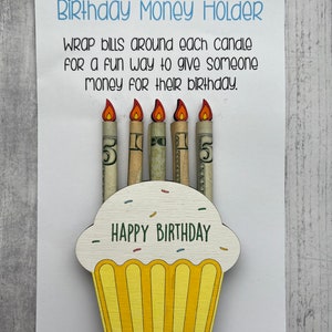 May include: A wooden cupcake money holder with the words "Happy Birthday" on the front. The cupcake has five candles, each wrapped with a dollar bill.