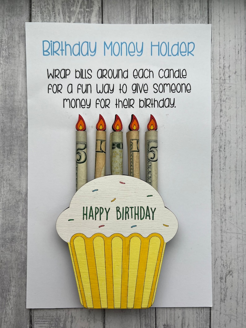 May include: A wooden cupcake money holder with the words "Happy Birthday" on the front. The cupcake has five candles, each wrapped with a dollar bill.