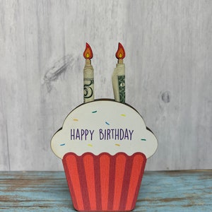 May include: Wooden cupcake money holder with red and white frosting and the words "Happy Birthday" on the front. Two dollar bills are inserted into the top of the cupcake as candles.