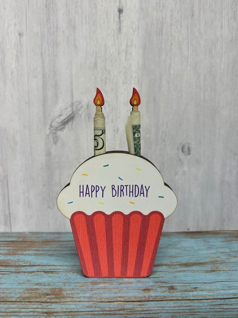 May include: Wooden cupcake money holder with red and white frosting and the words "Happy Birthday" on the front. Two dollar bills are inserted into the top of the cupcake as candles.