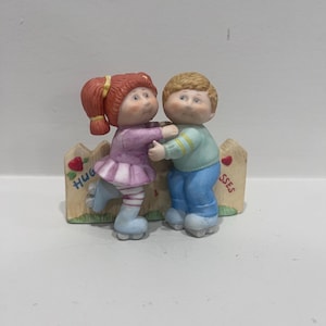 May include: A ceramic figurine of two children on roller skates leaning against a wooden fence. The girl has red hair in pigtails, wearing a purple top and striped leggings. The boy has blonde hair, wearing a light blue sweater and blue pants. The fence features red hearts and the text HUG SSES.