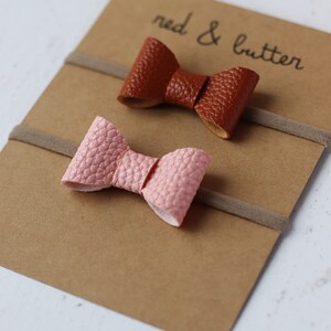 May include: Two sets of hair ties with bows. One bow is brown leather and the other is pink leather. The hair ties are on a brown card with the text "red & butter" written on it.