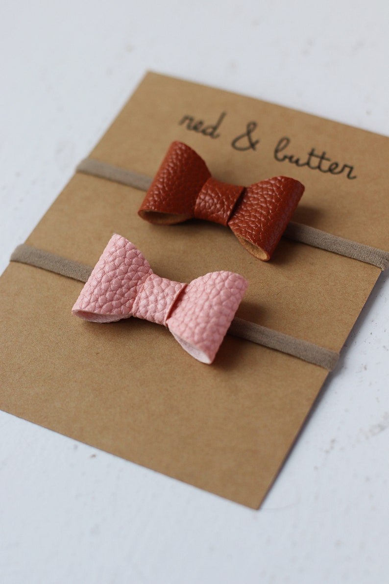 May include: Two sets of hair ties with bows. One bow is brown leather and the other is pink leather. The hair ties are on a brown card with the text "red & butter" written on it.