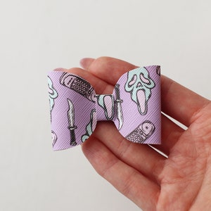 May include: A purple bow with a white and black print of a ghost face, knives, and a phone. The bow is made of fabric and is tied in a knot.