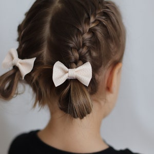 May include: A young girl with brown hair styled in two French braids with white bows at the ends.