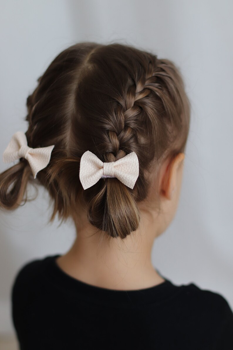 May include: A young girl with brown hair styled in two French braids with white bows at the ends.