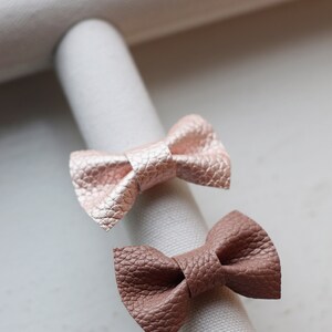 May include: Two leather hair bows, one in a light pink color and the other in a brown color, are displayed on a white fabric surface.