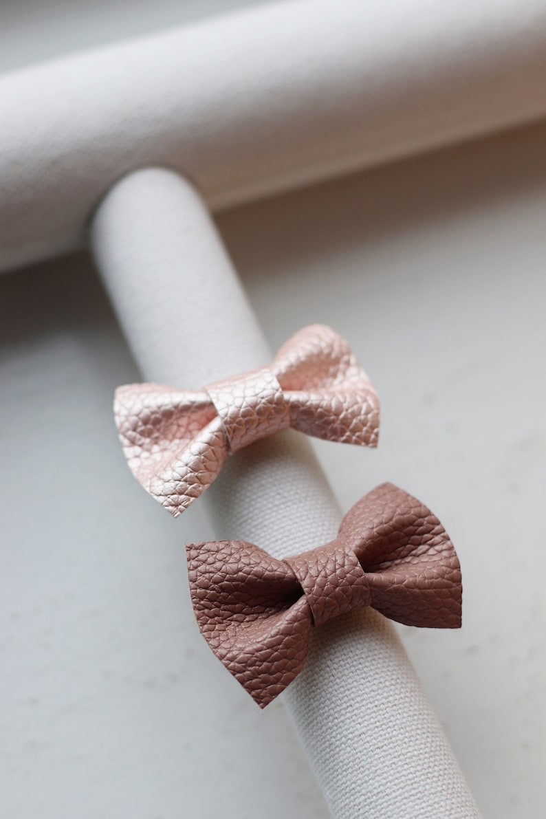 May include: Two leather hair bows, one in a light pink color and the other in a brown color, are displayed on a white fabric surface.