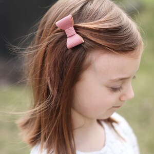 May include: A young girl with long brown hair wearing a white lace top with a small pink leather bow in her hair.