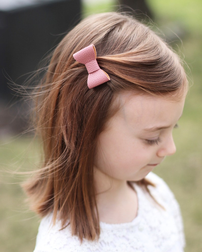 May include: A young girl with long brown hair wearing a white lace top with a small pink leather bow in her hair.