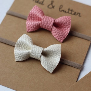 May include: Two fabric hair bows on brown cardstock with the text "red & butter". The bows are on nude elastic headbands. One bow is pink and the other is white.