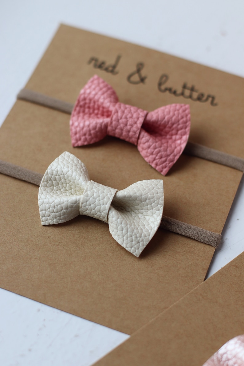 May include: Two fabric hair bows on brown cardstock with the text "red & butter". The bows are on nude elastic headbands. One bow is pink and the other is white.