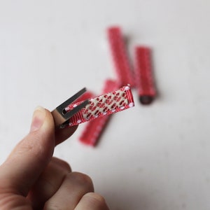 May include: A close-up of a red hair clip with white polka dots. The clip has a metal spring and a textured, beige fabric covering the top.