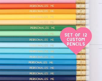May include: A set of 12 pencils in a rainbow of colors, each featuring a gold metal ferrule, a white eraser, and the gold-stamped text "PERSONALIZE ME". A pink heart-shaped graphic overlay displays the text "SET OF 12 CUSTOM PENCILS".
