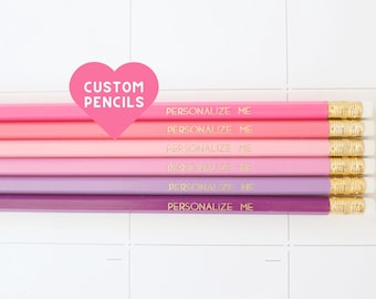 May include: A set of six wooden pencils in a gradient of pink and purple shades, each featuring a gold-toned metal ferrule with a white eraser and the gold-stamped text "PERSONALIZE ME". A pink heart graphic with the text "CUSTOM PENCILS" is overlaid.