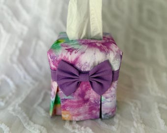 May include: A fabric tissue box cover with a tie-dye pattern in shades of purple, pink, green, and yellow. A purple bow is attached to the front. White tissues are visible at the top.