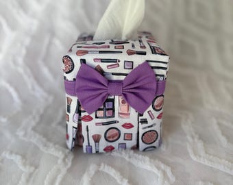 May include: A square tissue box cover with a white background and a pattern of makeup items in pink, purple, and black. A purple bow is attached to the front. White tissues are coming out of the top.